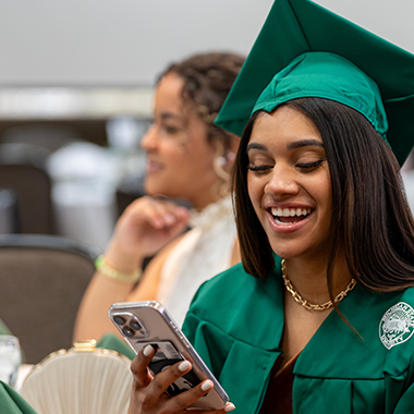 Female student wearing graduation hat and gown, smiling while looking at her phone.